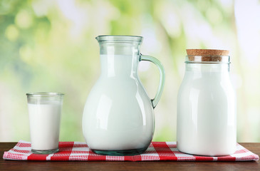 Pitcher, jar and glass of milk on wooden table, on nature background