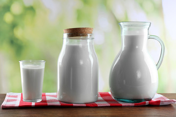 Pitcher, jar and glass of milk on wooden table, on nature background
