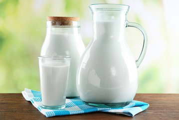Pitcher, jar and glass of milk on wooden table, on nature background