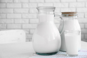 Pitcher, jar and glass of milk on wooden table, on bricks wall background