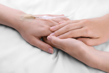 Obraz na płótnie Canvas Female hands holding patient hand with dropper needle on bed close-up