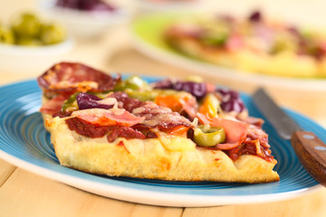Fresh homemade pizza piece with tomato sauce, ham, salami, olives, bell pepper and cheese on top, served on blue plate with knife (Selective Focus, Focus on the front of the green olive slice)