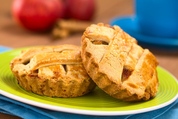 Small round apple pie with lattice crust on green plate, with a blue cup, apples and cinnamon sticks in the back  (Selective Focus, Focus on the front of the left pie and one third into the right pie)