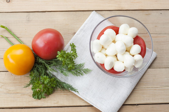 Mozzarella cheese in a glass bowl, tomatoes, sliced tomatoes and herbs on a wooden table, selective focus