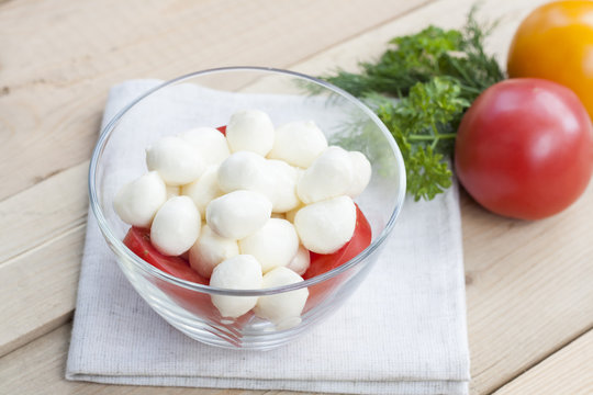 Mozzarella cheese in a glass bowl, tomatoes, sliced tomatoes and herbs on a wooden table, selective focus