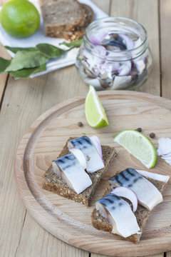 Slices of marinated mackerel with onion in a jar, lime, laurel and bread on wooden board, selective focus
