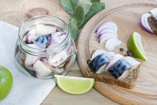 Slices of marinated mackerel with onion in a jar, lime, laurel and bread on wooden board, selective focus