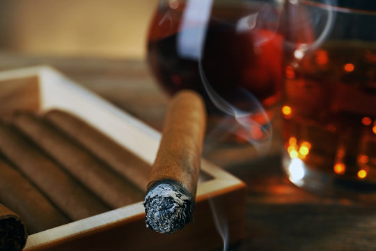 Cigars and burning one with cognac on wooden table, closeup