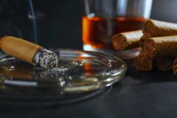 Group of cigars and burning one with whiskey on table, closeup