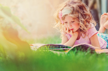 adorable curly child girl reading book and dreaming on summer backyard