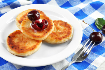 Fritters of cottage cheese with cherry and cream in plate on checkered napkin, closeup