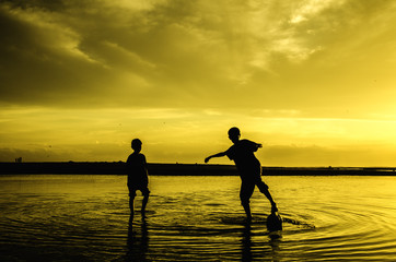 image silhouette of two boys play beach soccer during sunset sunrise. blur on water and sky. reflect on water