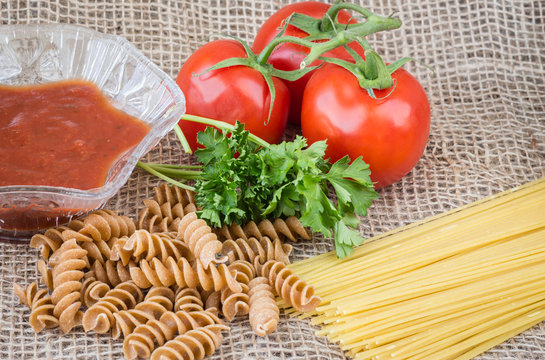 horizontal image of raw spaghetti and whole wheat pasta with tomato on the vine and a dish of salsa on burlap background. 