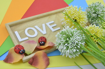 white word and love symbol on colourfull background.green, yellow and white artificial flower placed on right.petal placed on bottom