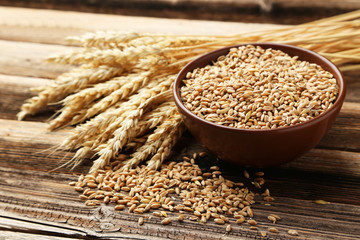 Ears of wheat and bowl of wheat grains on brown wooden backgroun