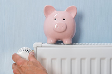 Man Holding Thermostat With Piggy Bank On Radiator