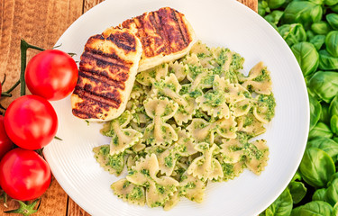 Top view of grilled halloumi cheese with pesto farfalle near a basil bed  and a vine of tomatoes on a wooden table.