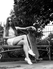 Obraz premium Beautiful girl with longboard sits on bench in front of green