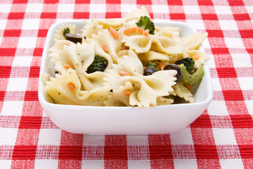 Bowtie Pasta Salad – Bowtie (farfalle) pasta salad with black olives, broccoli and carrots. On a red checked background.