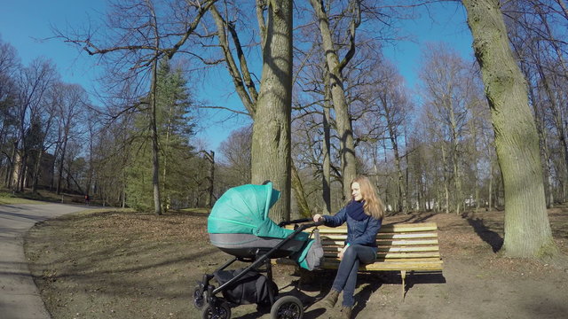 Happy smiling young mother woman with baby in buggy sitting on bench under tree in spring park. Healthy leisure in fresh air. Wide angle shot. 4K UHD.
