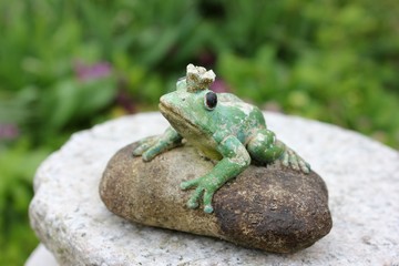 Waiting weathered stone frog statue waiting on a stone to be kissed in the garden, seen from birds eyes view looking up. Character from a Russian fairytale. Black eyes. Blurry green background.