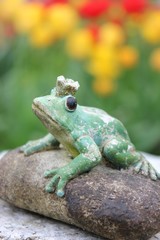 Waiting weathered stone frog statue waiting on a stone to be kissed in the garden, seen from front view. Character from a Russian fairytale. Black eyes. Blurry tulip flower garden background, green.