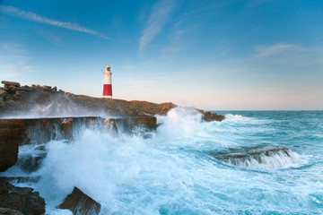 Stormy seas crash over the rocks with the lighthouse in the background at Portland Bill in...
