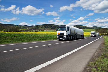 Fototapeta na wymiar White tanker and trucks driving along the asphalt road around the yellow flowering rapeseed field