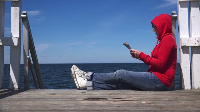 Young woman reading fanzine magazine at the edge of wooden ocean pier.