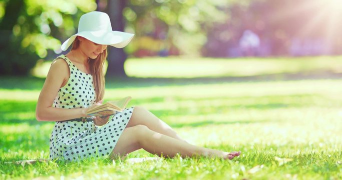 Woman reading a book in the park outdoors. Slow motion. Dolly shot. Beautiful romantic lady in white hat sitting on the green grass lawn and looking through the novel pages and daydreaming. 4K, DCi.