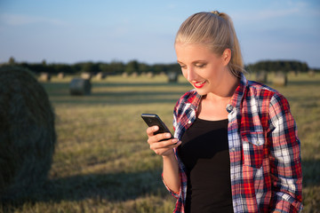 beautiful blonde woman with mobile phone in field with haystacks