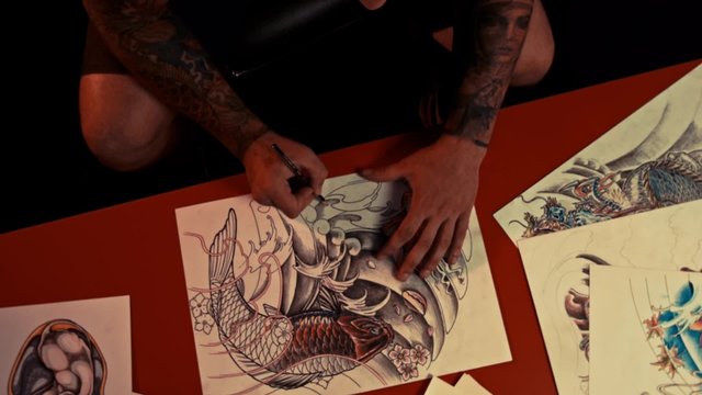 Seated tattoo-artist in spectacles draws sketch on the table