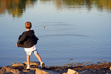 Young Boy Skips Stones – A young boy skips throws a stone into the lake, skipping it across the water and creating ripples. Early evening light.
