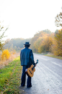 Lonely guitarist looking at empty country road in autumn.