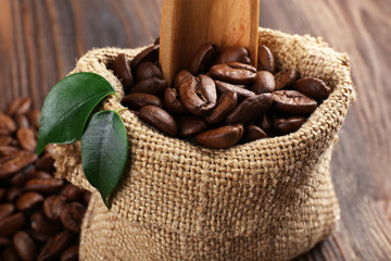 Coffee beans with leaves and spoon in bag on wooden table close up