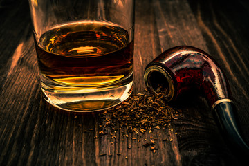 Glass of whiskey and tobacco pipe with tobacco leaves are scattered on the wooden table