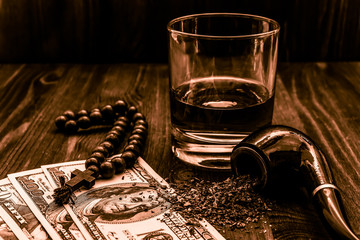 Glass of whiskey and rosary with cross and a money with tobacco leaves on a wooden table