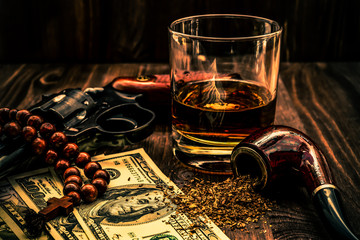 Glass of whiskey and revolver with a rosary with cross and tobacco leaves on a wooden table © Georgii Shipin