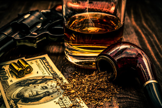 Glass of whiskey and revolver with a money and tobacco pipe on a wooden table