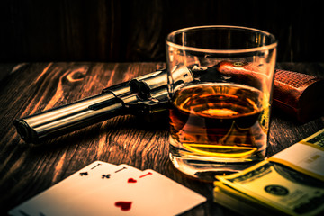Glass of whiskey and playing cards with revolver and money on the wooden table