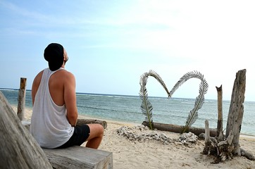 A single man sits alone on the beach in front of a big heart. He looks up to the sky. Perhaps he is sad, depressed and alone. Perhaps he just waits for his girlfriend. 