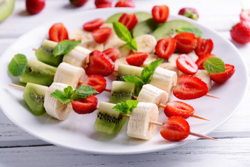 Fresh fruits on skewers in plate on wooden table, closeup