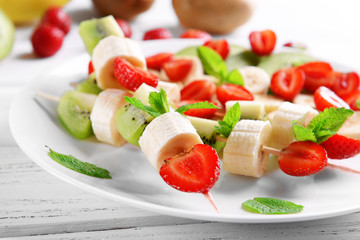 Fresh fruits on skewers in plate on wooden table, closeup