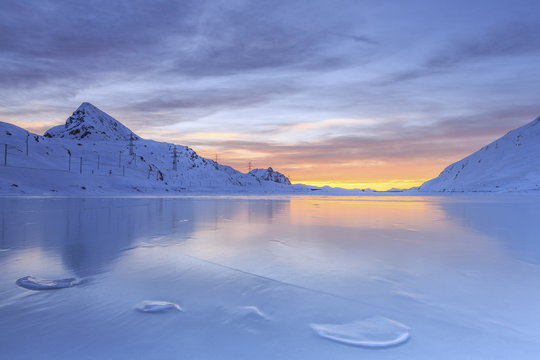 The colors of dawn invading the smooth surface of Lago Bianco exceptionally icy, Bernina Pass, Graubunden, Swiss Alps