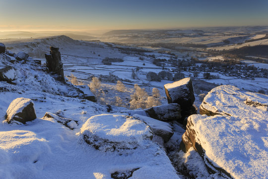 Snow on millstone lit by sunrise, Curbar and Baslow Edge with misty hills and wintry trees, Peak District, Derbyshire