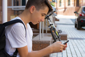 Outside portrait of teen boy. Handsome teenager carrying backpack on one shoulder and smiling, communicating by phone