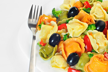 Salad made with tortellini, olives, broccoli, red pepper, on a plate with a fork, white background