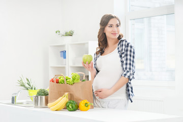 Beautiful pregnant woman in the kitchen with shopping bag and apple.