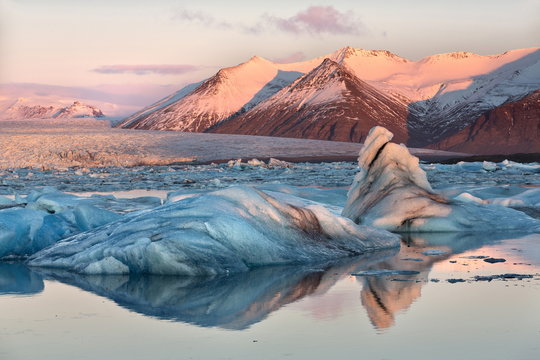 View across the calm water of Jokulsarlon glacial lagoon towards snow-capped mountains and icebergs bathed in winter morning light, at the head of the Breidamerkurjokull Glacier on the edge of the Vatnajokull National Park, South Iceland, Iceland, Polar R