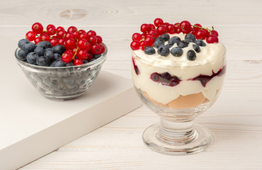 Sweet dessert in glass with biscuits, cream, currants and blueberries on wooden background.